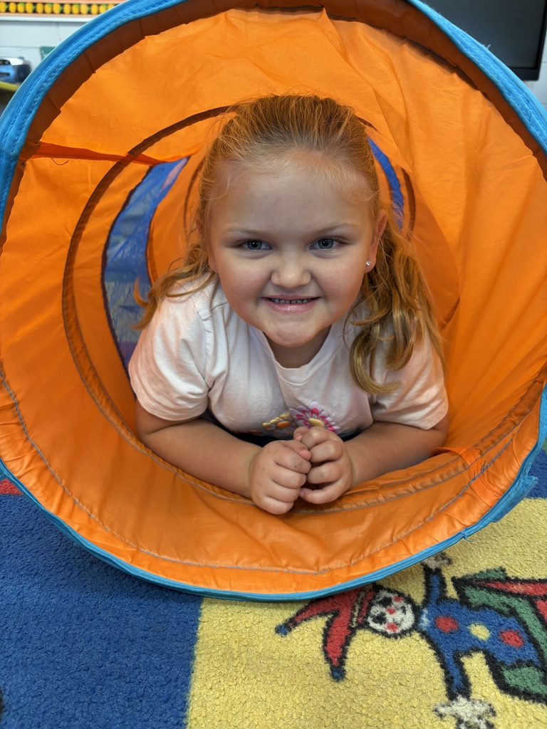 Young girl with blonde hair smiles and peers out of orange and blue tunnel on a multicolored floor.