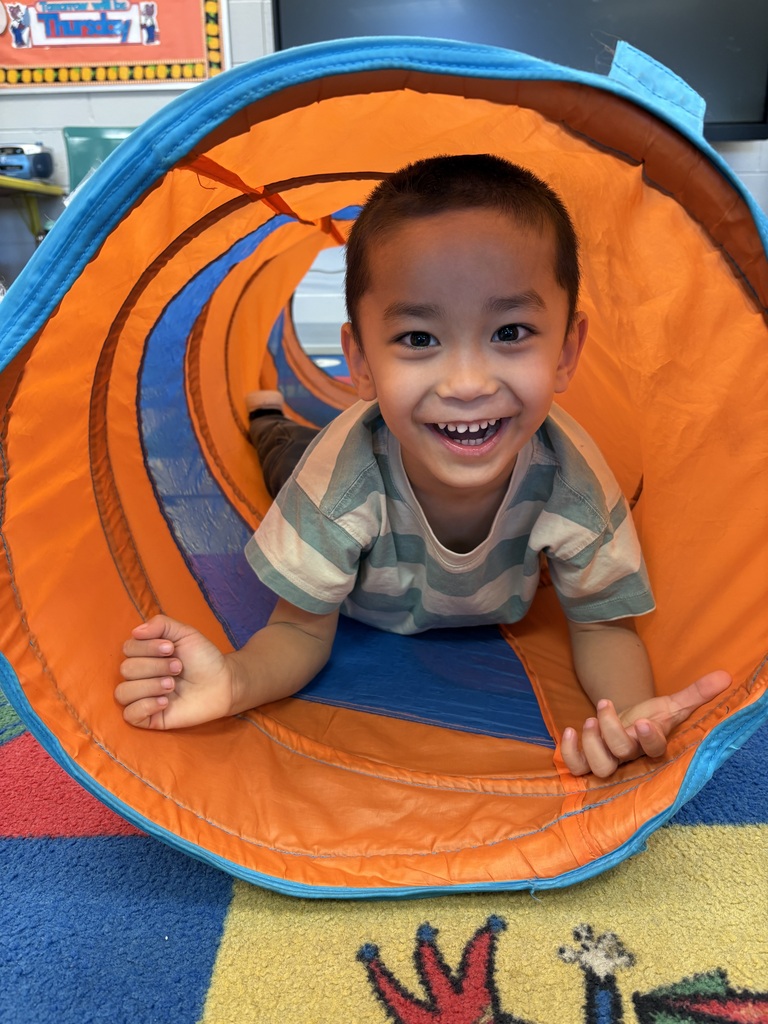Child with a striped shirt smiling inside an orange and blue tunnel, playing on a colorful floor.
