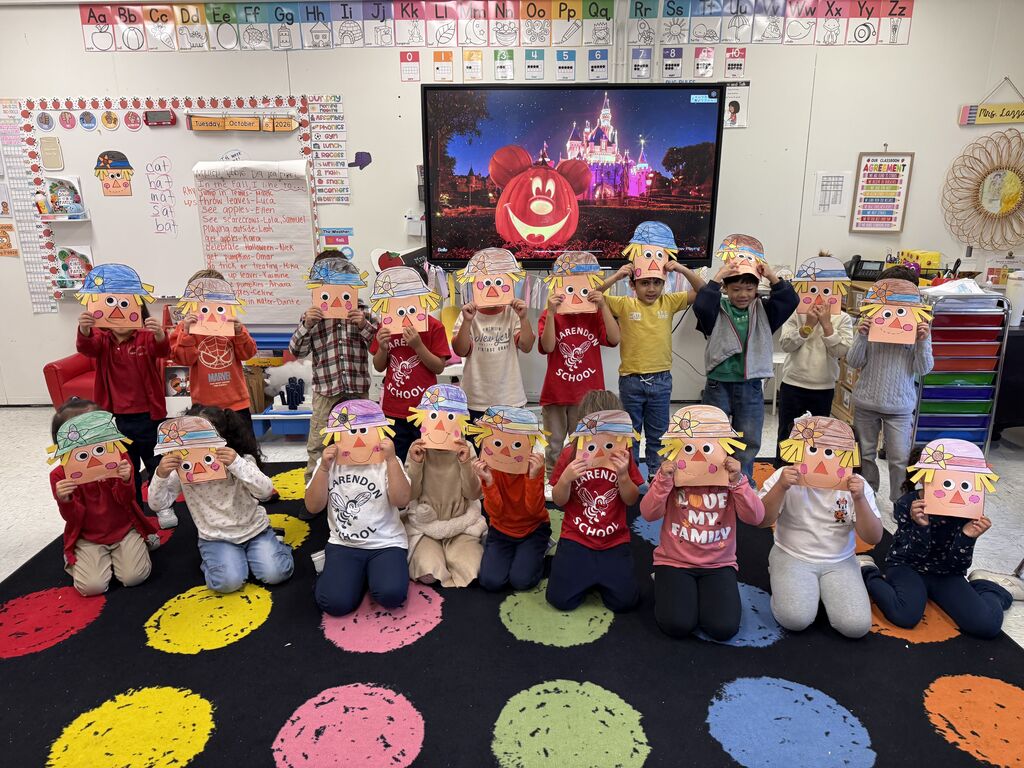 A classroom of students wearing paper masks sit in a circle on the floor. A large screen shows a movie.