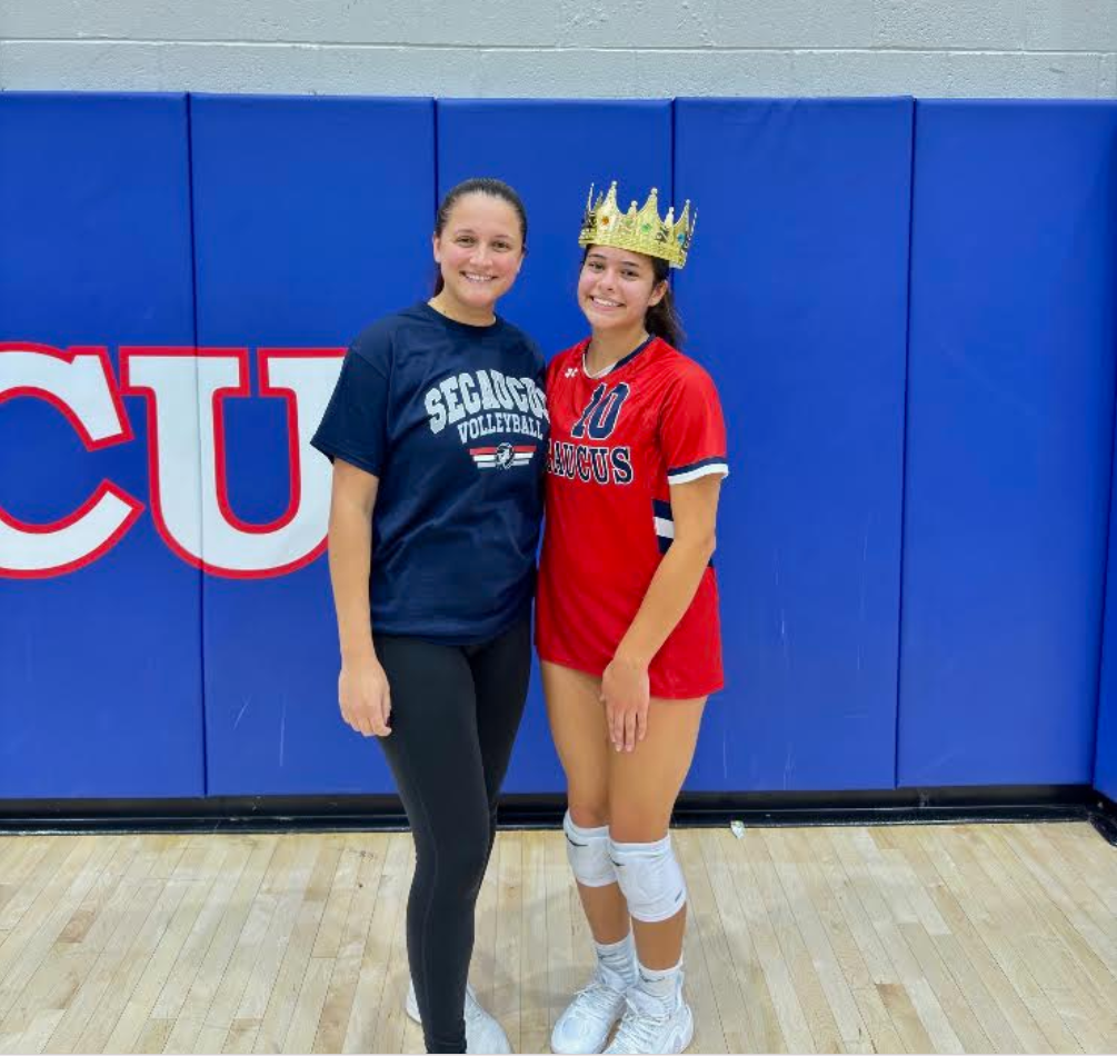 Two women standing on a basketball court, one wearing a crown. A blue wall with white letters reads "CU".