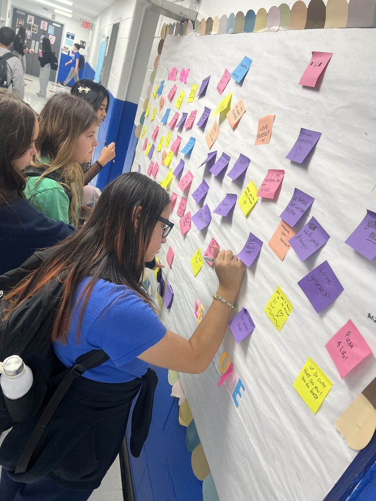 Students contribute to a bulletin board covered with colorful sticky notes. A woman with long hair in a blue shirt writes on the board.