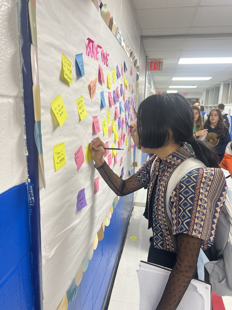 A person stands at a wall with sticky notes, writing on a yellow note. The wall is white and blue.