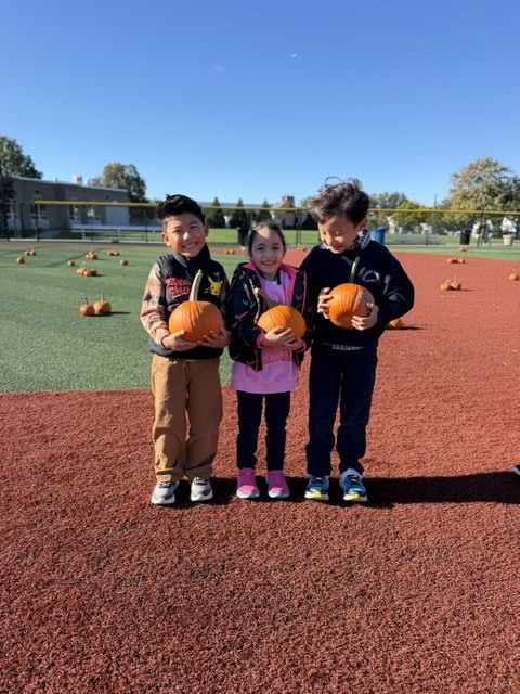 Three children stand in a pumpkin patch, each holding a pumpkin. They wear jackets and sneakers. Behind them, a field with numerous pumpkins and trees is visible.