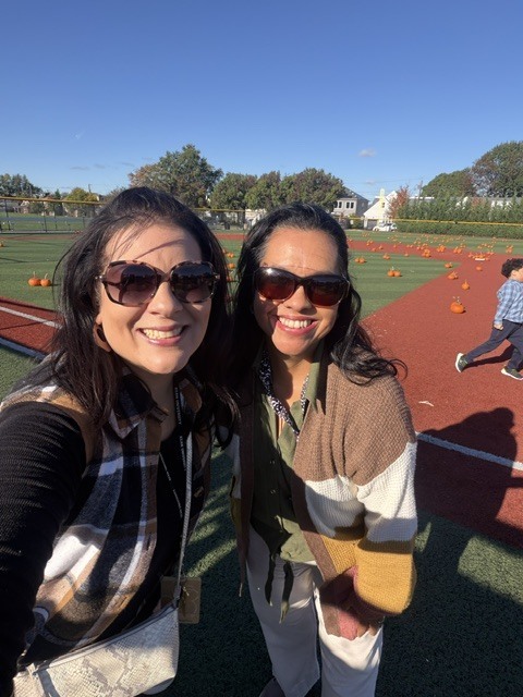 Two women with sunglasses smile for a selfie on a pumpkin-filled field. A child runs in the background.