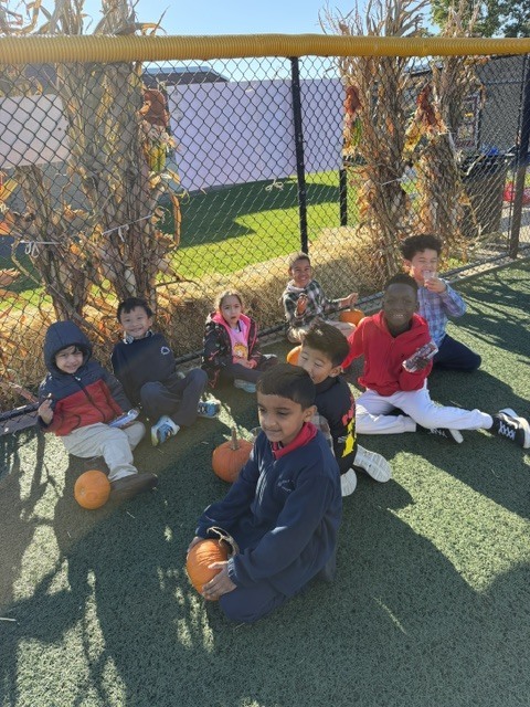 Several children sitting on a green play surface, holding pumpkins, with a chain-link fence behind them.