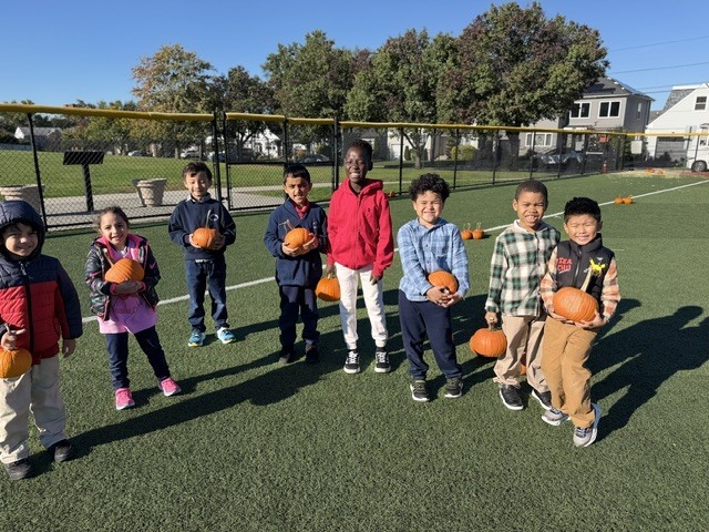 Nine children stand on a field, holding pumpkins. They wear various coats and sneakers. A fence and trees surround the area.