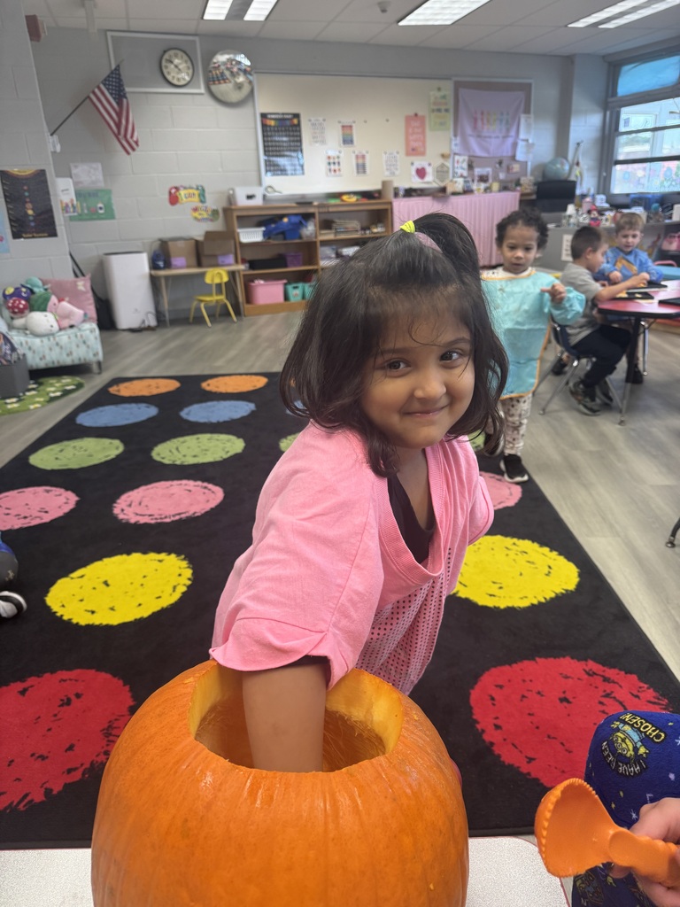 Girl with pigtails inside pumpkin; colorful dots on floor; children seated at tables; classroom with shelves, flag, and clock.
