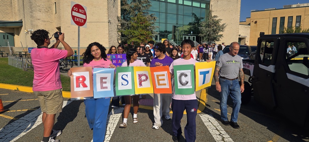 Group of students carrying a multi-colored banner with letters forming the word "RESPECT" walking in a crosswalk.