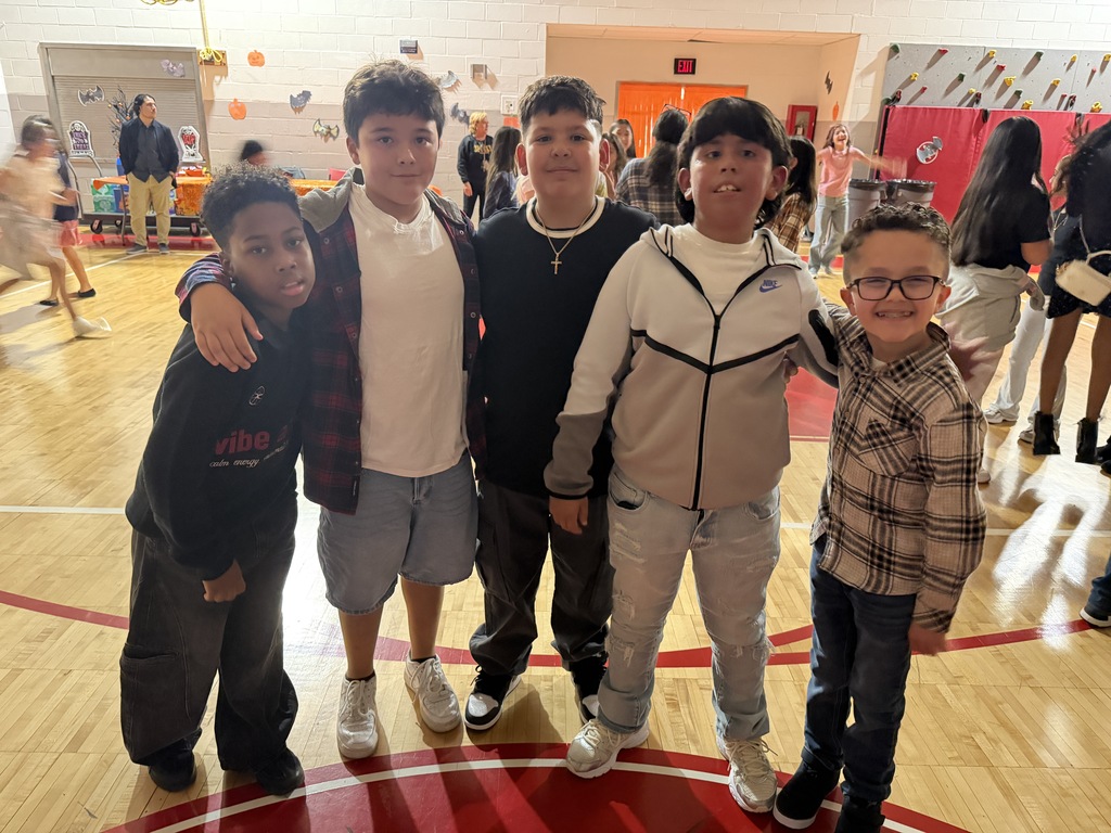 Five young boys standing on a basketball court with others walking in the background.
