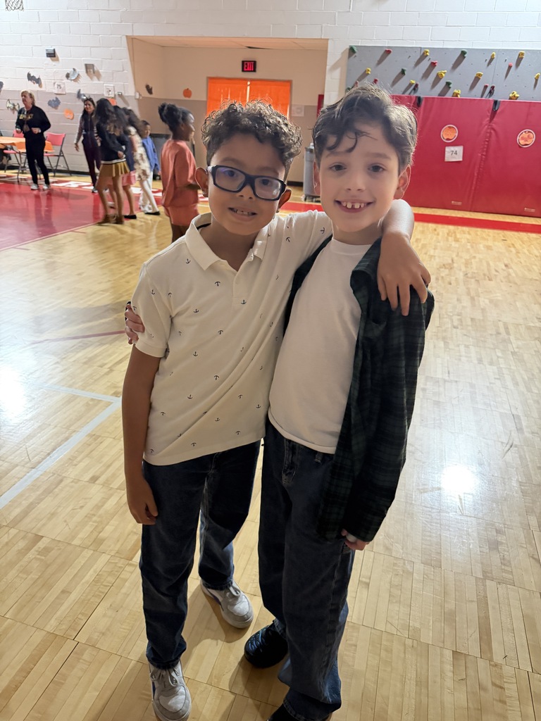 Two boys stand on a gym floor, one wearing glasses and both smiling for the camera.