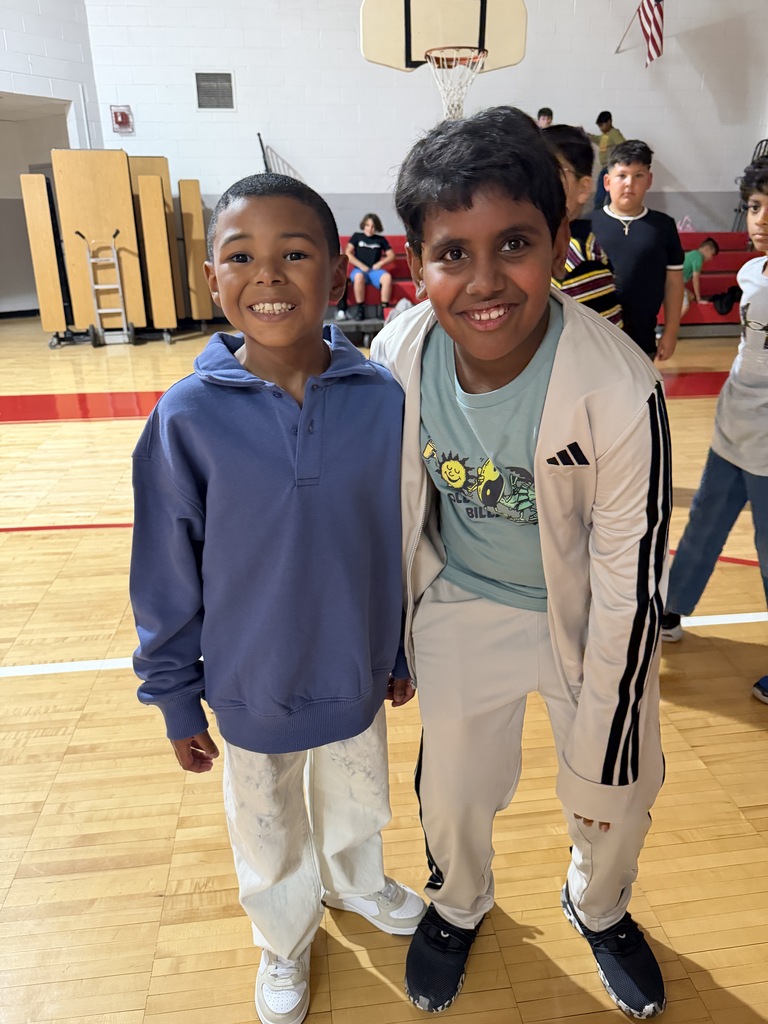 Two boys smile for photo in a gym; one wears a blue hoodie and white pants, the other a white jacket and pants.