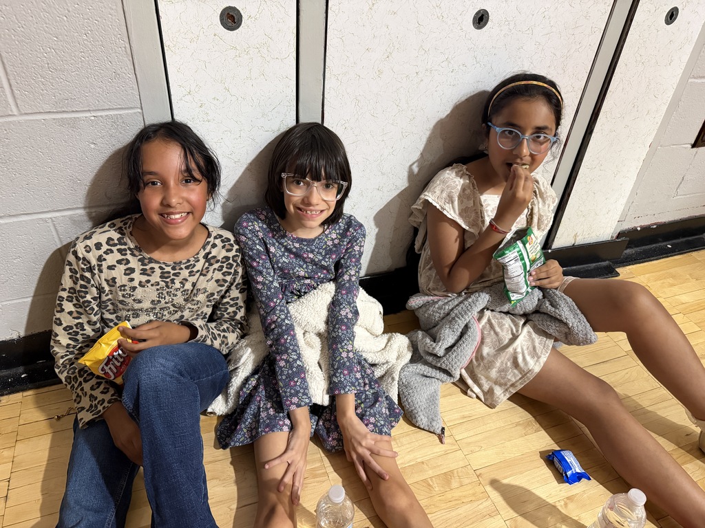 Three girls sit on the floor. Two hold snacks. One holds a drink. A white wall is behind them.