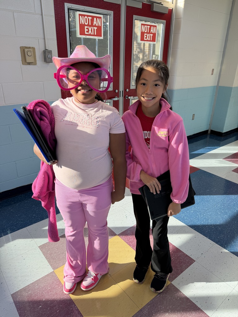 Two girls in front of double doors, one holding books and wearing glasses and a hat, other wearing pink.