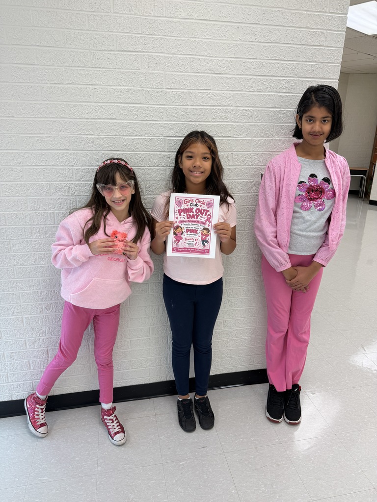 Three young girls in pink clothes stand close together. The girl in the middle holds a sign, while the others hold small pink items.