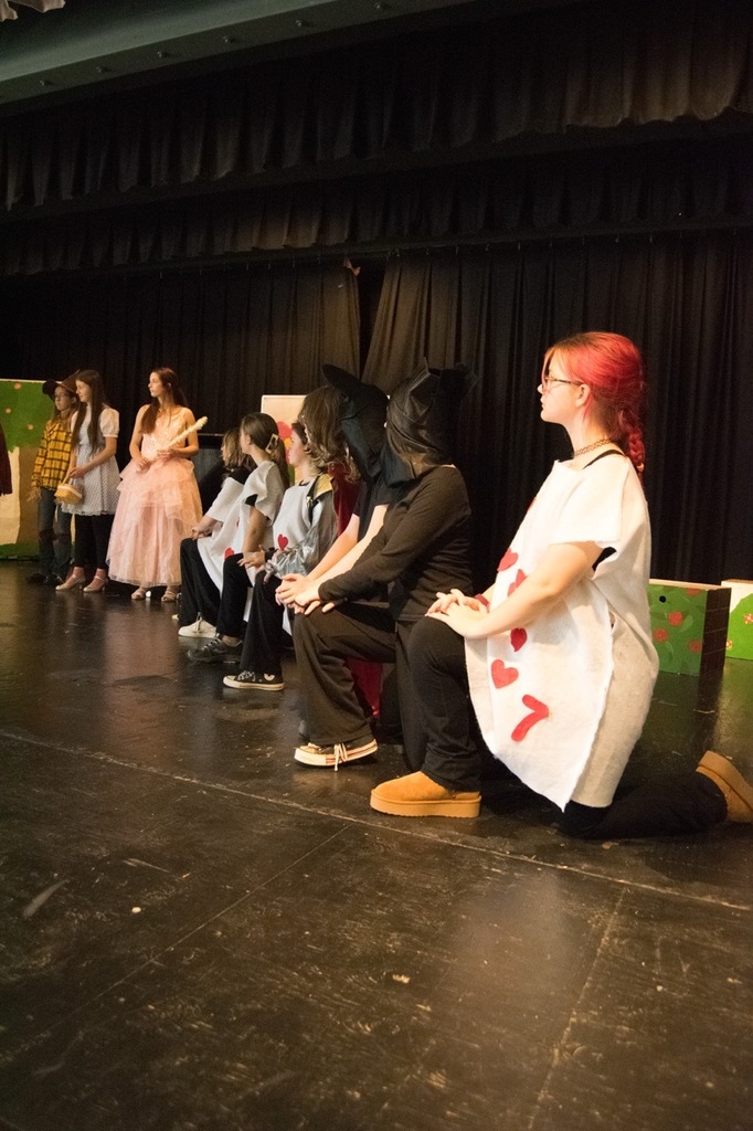 Cascade Middle School drama students in costume rehearsing for “Dorothy in Wonderland,” with characters from The Wizard of Oz and Alice in Wonderland gathered on stage under colorful lights.
