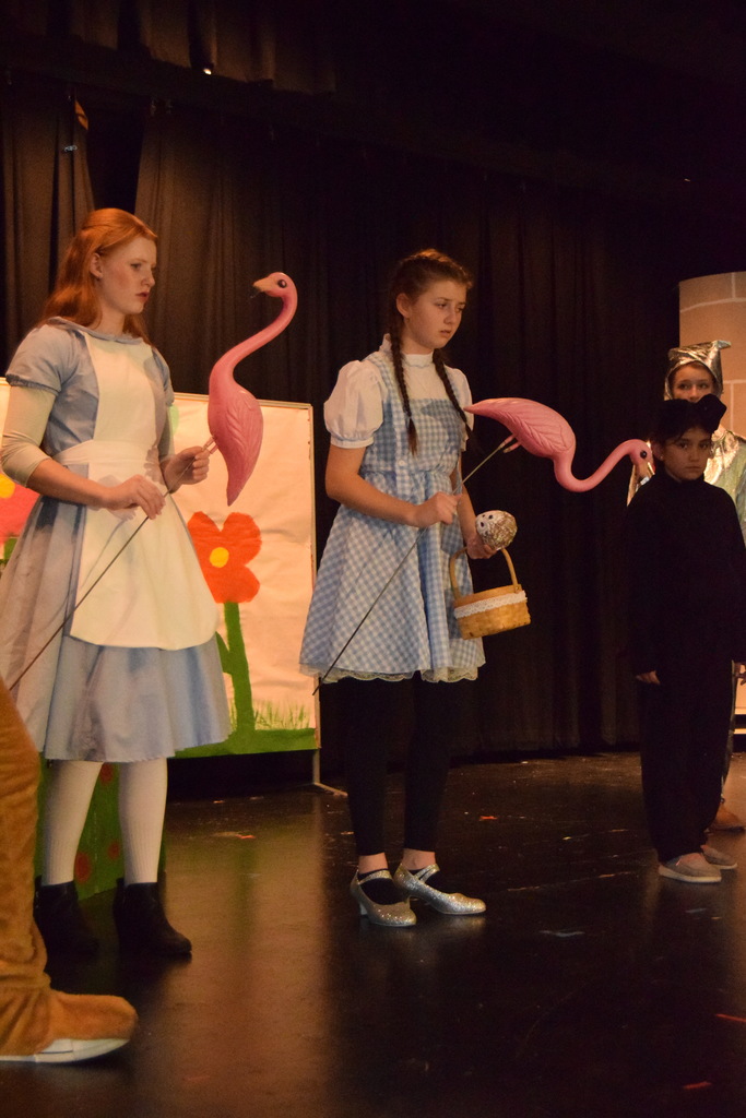 Cascade Middle School drama students in costume rehearsing for “Dorothy in Wonderland,” with characters from The Wizard of Oz and Alice in Wonderland gathered on stage under colorful lights.