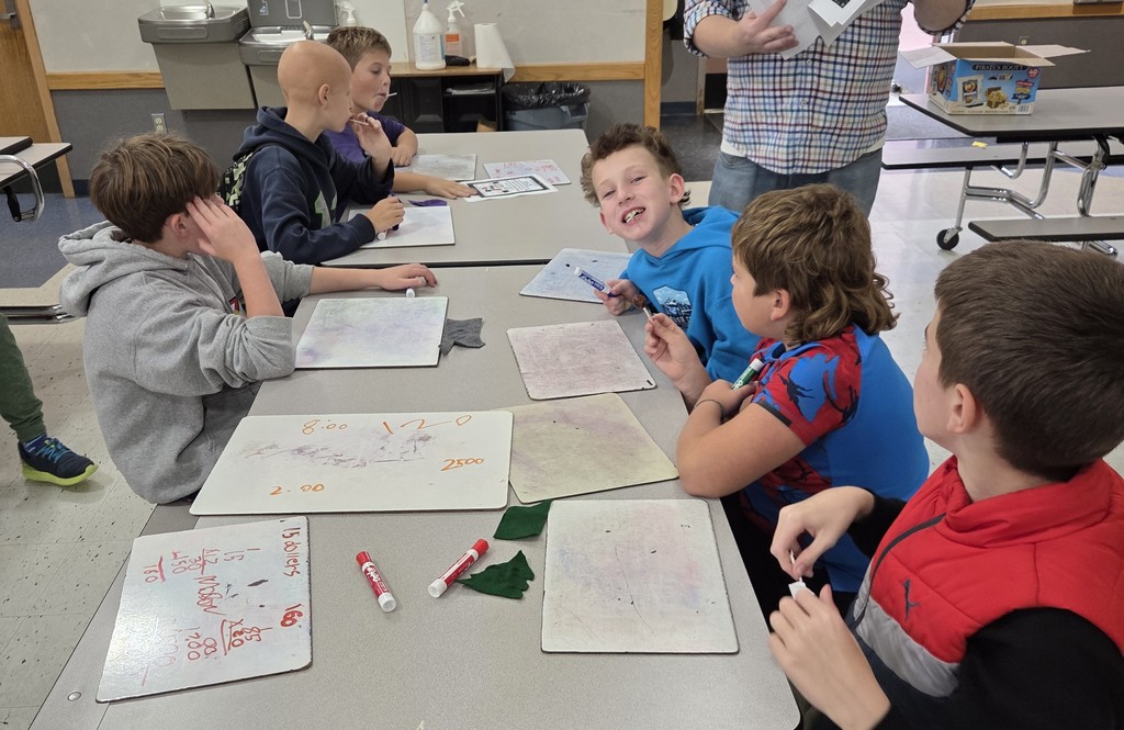 Students gather around a table at Big Lake Elementary, smiling as they work together on a math problem during the first Math Olympiad meeting.