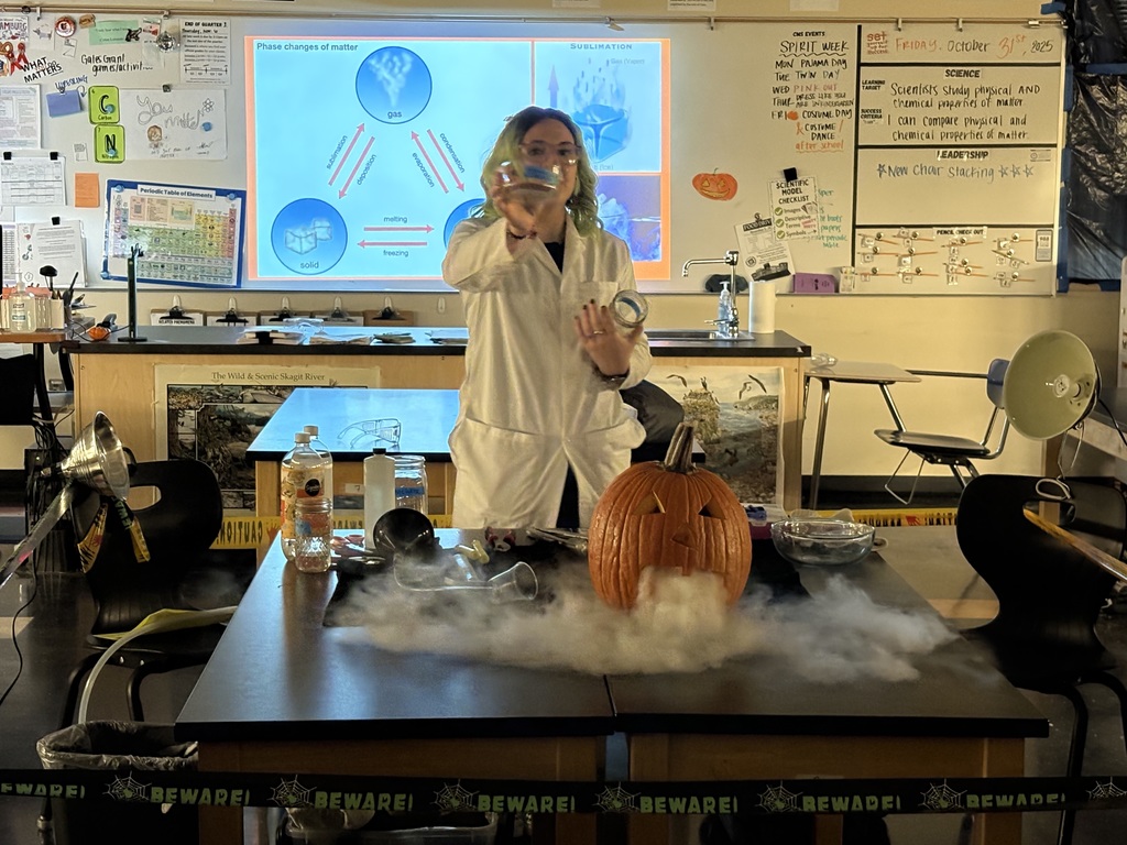 Science teacher Heather Hirschi, dressed as a mad scientist, performs glowing dry ice and highlighter ink demonstrations for Cascade Middle School students. Under black lights, fog, bubbles, and bright colors show sublimation and gas reactions while students watch safely from their desks.