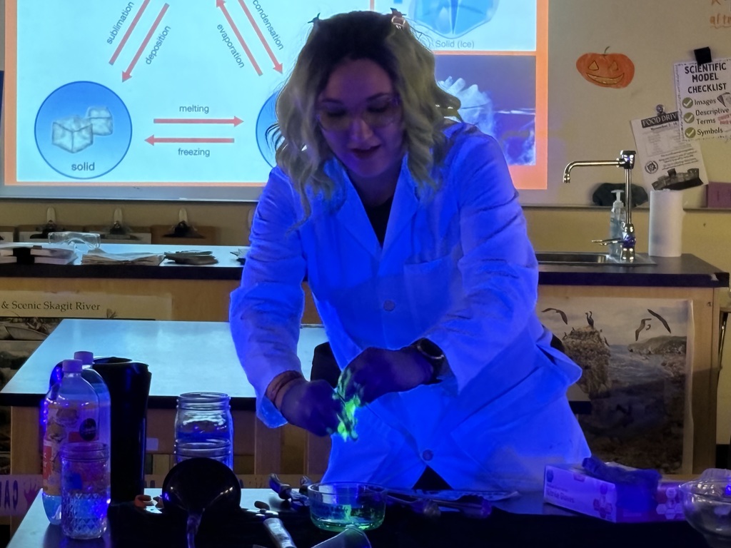 Science teacher Heather Hirschi, dressed as a mad scientist, performs glowing dry ice and highlighter ink demonstrations for Cascade Middle School students. Under black lights, fog, bubbles, and bright colors show sublimation and gas reactions while students watch safely from their desks.