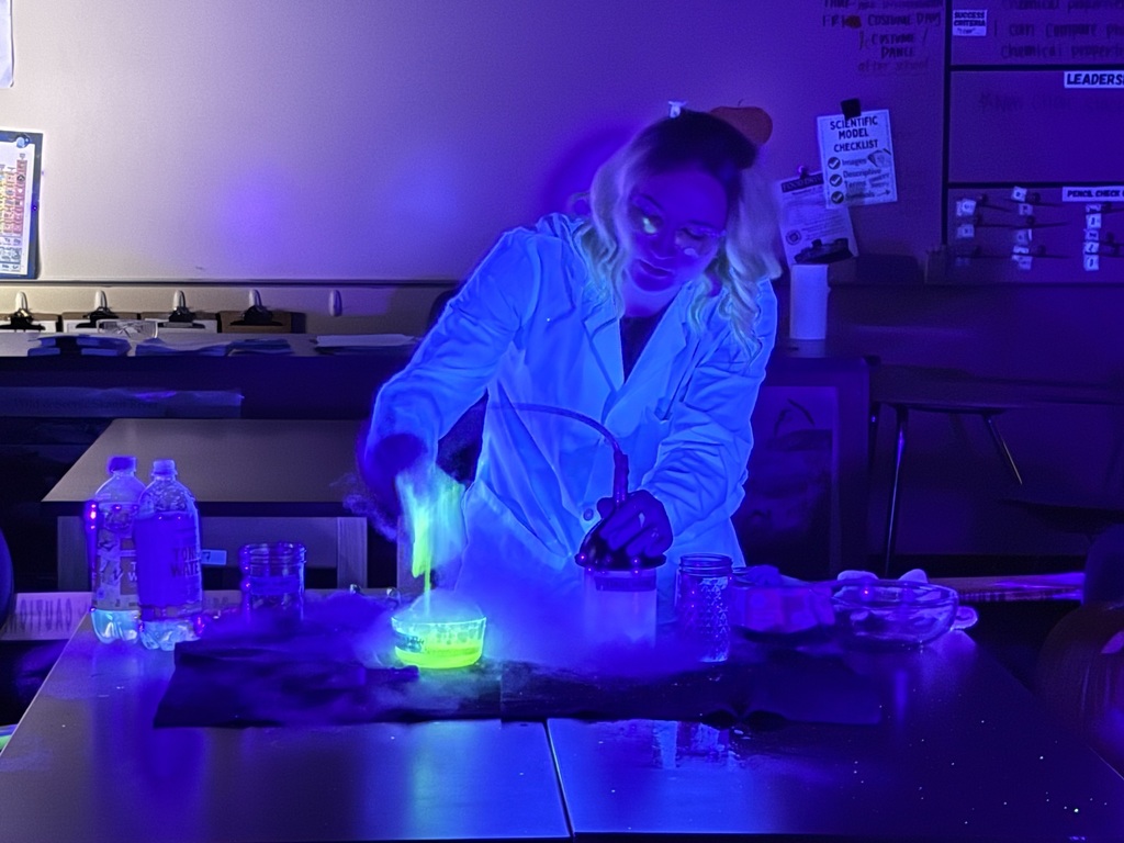 Science teacher Heather Hirschi, dressed as a mad scientist, performs glowing dry ice and highlighter ink demonstrations for Cascade Middle School students. Under black lights, fog, bubbles, and bright colors show sublimation and gas reactions while students watch safely from their desks.