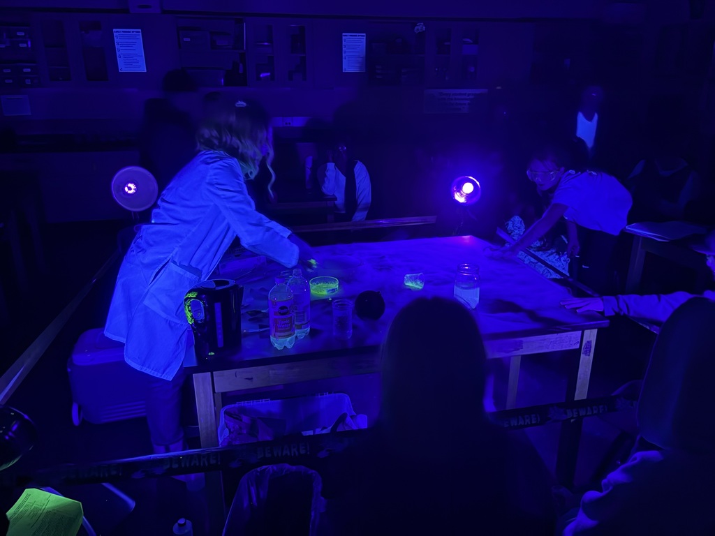 Science teacher Heather Hirschi, dressed as a mad scientist, performs glowing dry ice and highlighter ink demonstrations for Cascade Middle School students. Under black lights, fog, bubbles, and bright colors show sublimation and gas reactions while students watch safely from their desks.
