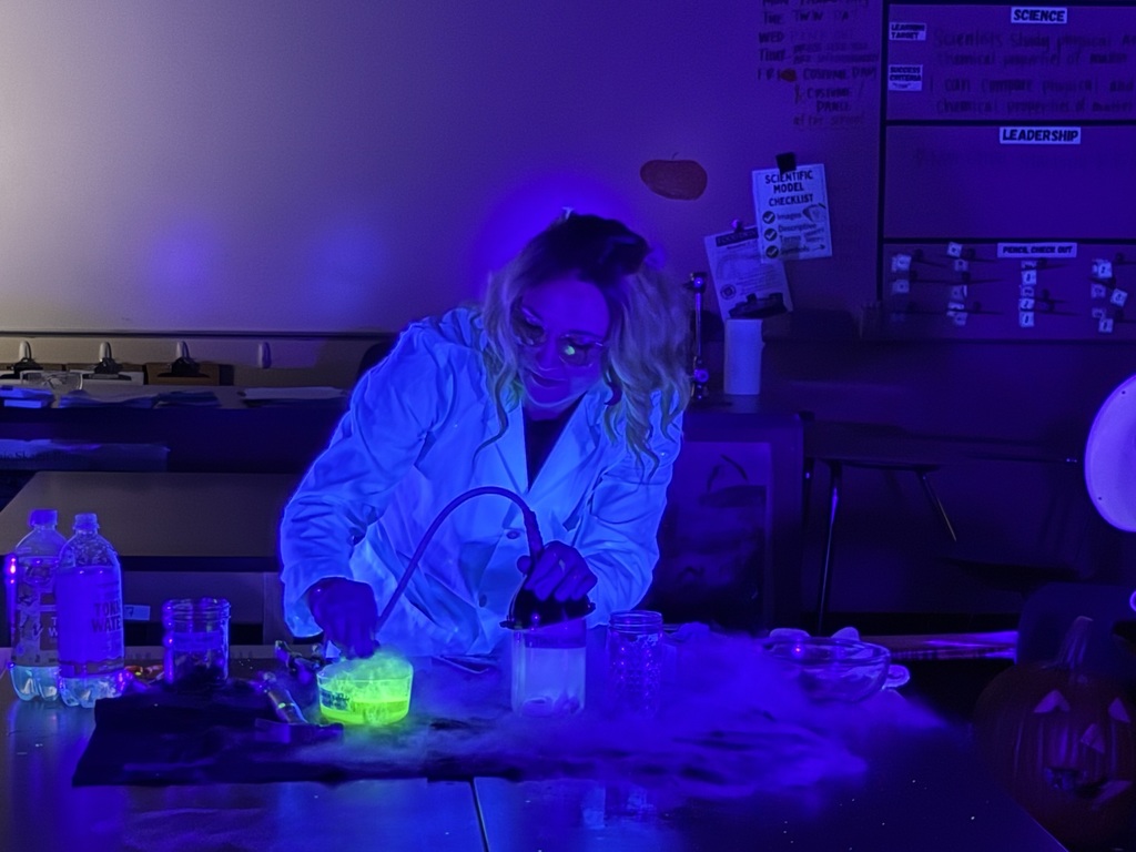 Science teacher Heather Hirschi, dressed as a mad scientist, performs glowing dry ice and highlighter ink demonstrations for Cascade Middle School students. Under black lights, fog, bubbles, and bright colors show sublimation and gas reactions while students watch safely from their desks.
