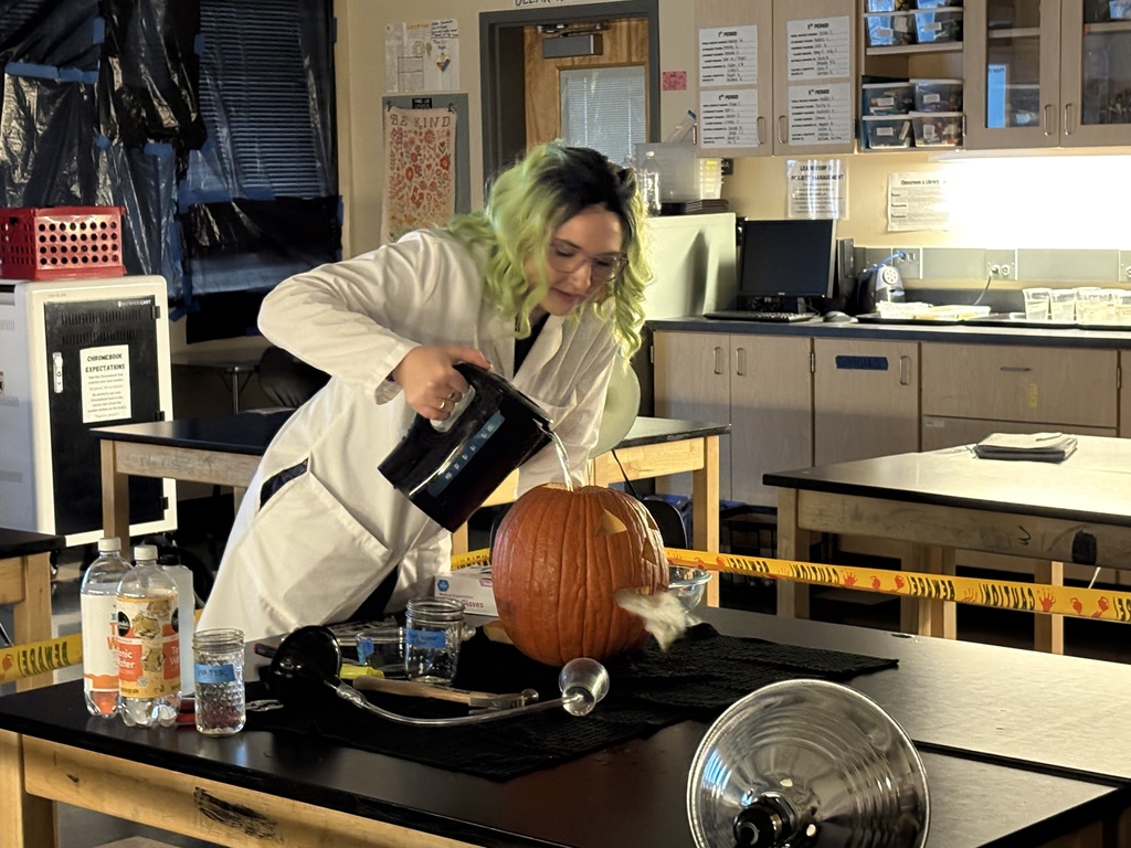Science teacher Heather Hirschi, dressed as a mad scientist, performs glowing dry ice and highlighter ink demonstrations for Cascade Middle School students. Under black lights, fog, bubbles, and bright colors show sublimation and gas reactions while students watch safely from their desks.