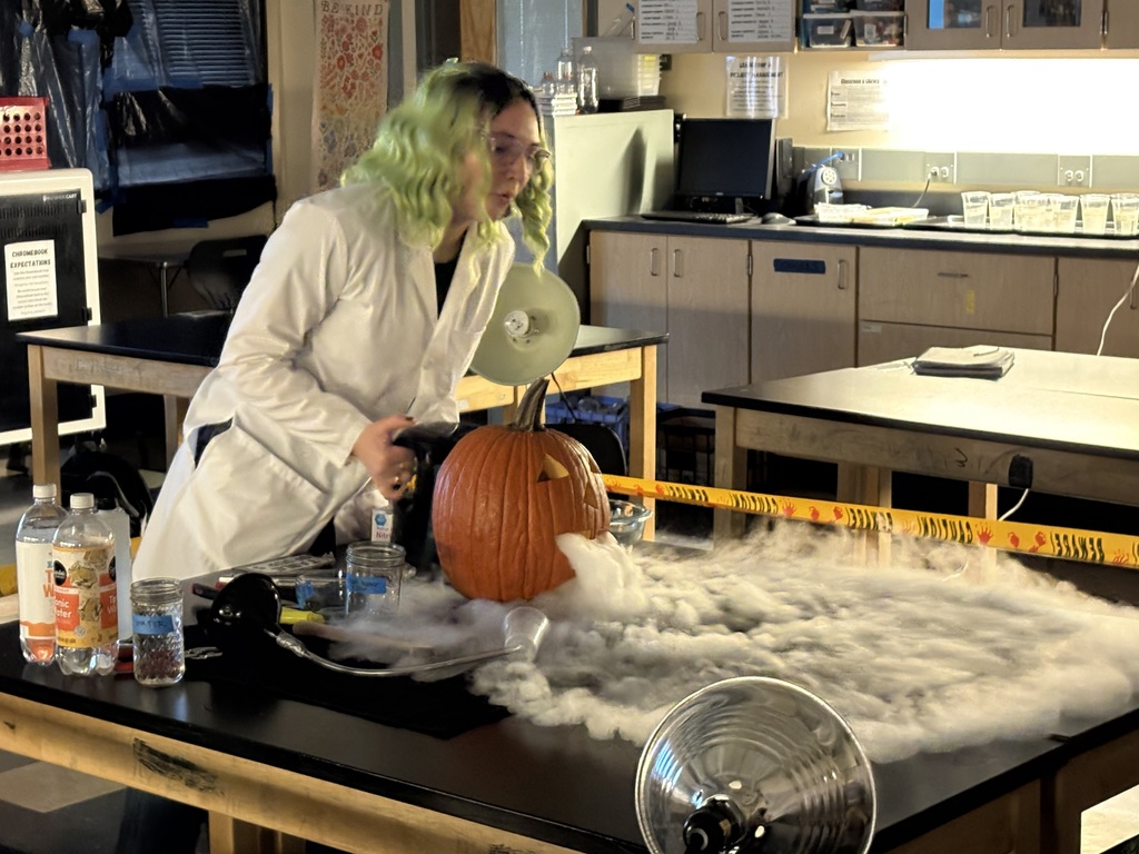 Science teacher Heather Hirschi, dressed as a mad scientist, performs glowing dry ice and highlighter ink demonstrations for Cascade Middle School students. Under black lights, fog, bubbles, and bright colors show sublimation and gas reactions while students watch safely from their desks.