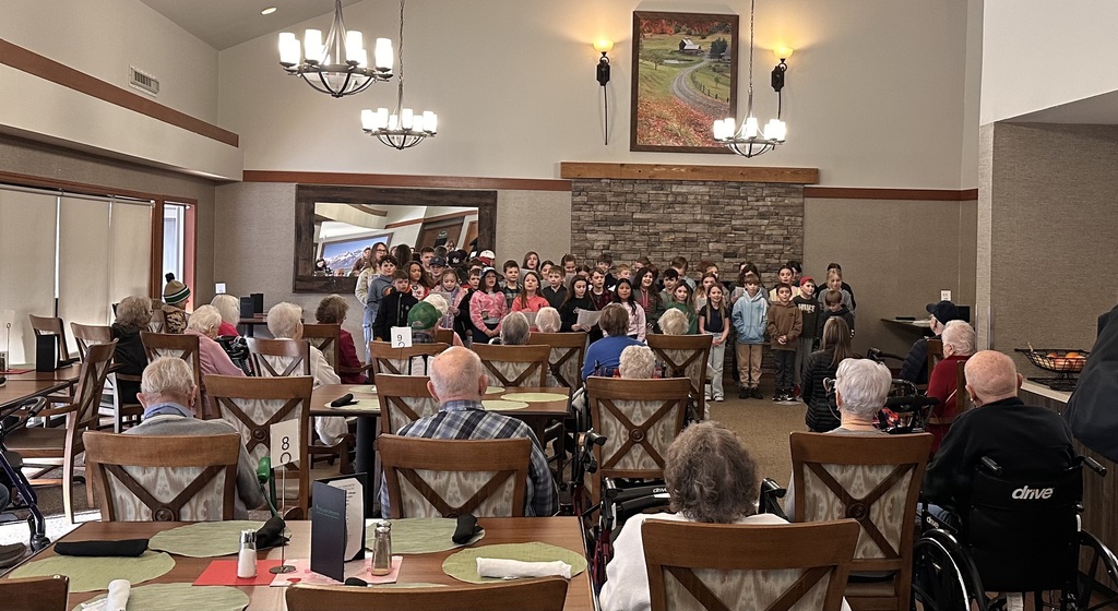 Clear Lake Elementary third and fifth grade students stand in a group performing a music program for senior residents at Country Meadow Village in Sedro-Woolley. Students sing and smile while residents watch from chairs and wheelchairs, creating a warm, intergenerational moment indoors.