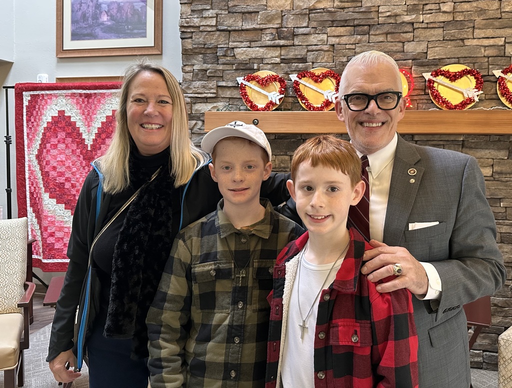Clear Lake Elementary third and fifth grade students stand in a group performing a music program for senior residents at Country Meadow Village in Sedro-Woolley. Students sing and smile while residents watch from chairs and wheelchairs, creating a warm, intergenerational moment indoors.