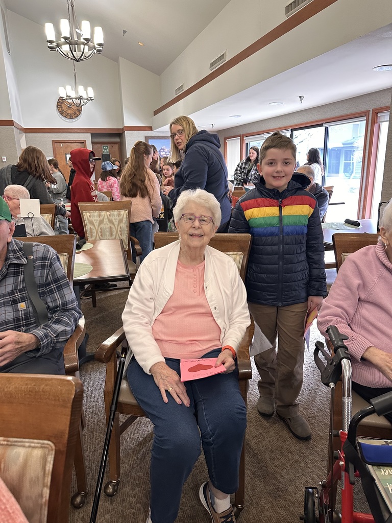 Clear Lake Elementary third and fifth grade students stand in a group performing a music program for senior residents at Country Meadow Village in Sedro-Woolley. Students sing and smile while residents watch from chairs and wheelchairs, creating a warm, intergenerational moment indoors.