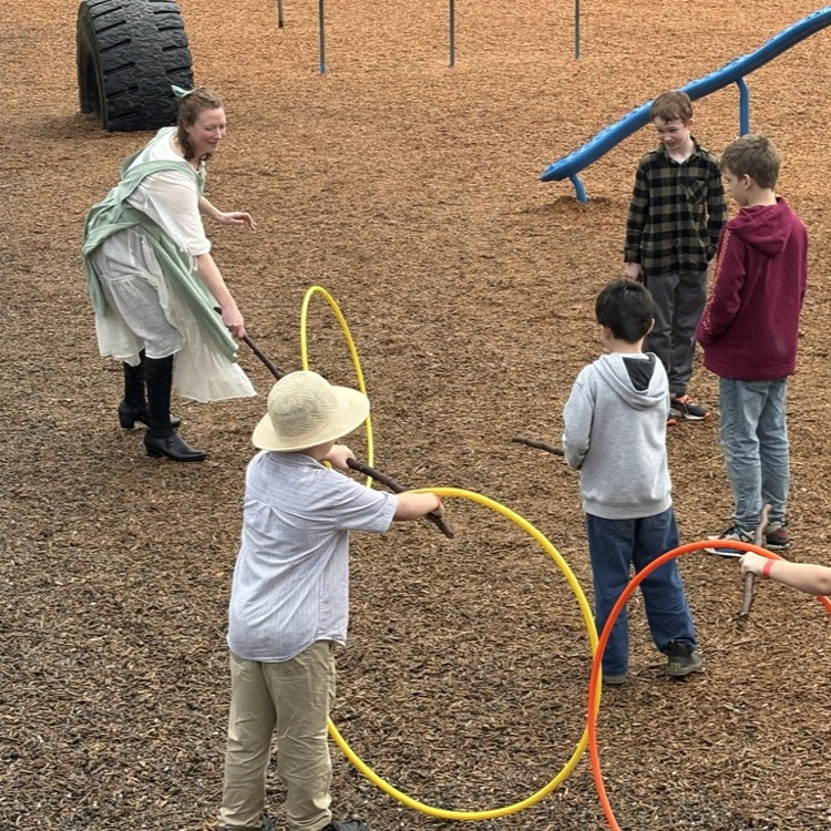 Student using a stick to roll a hoop during a pioneer-style game outside.