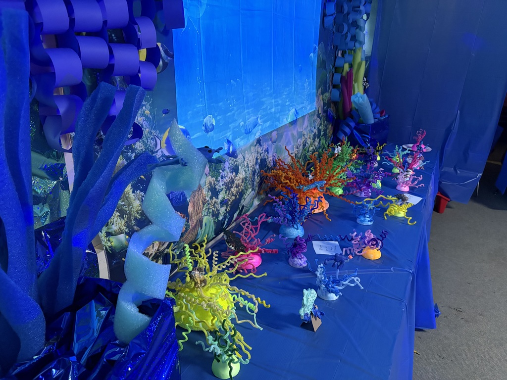 A classroom transformed into a large-scale ocean habitat, with blue fabric draped across the ceiling and handmade sea creatures hanging overhead, surrounding student-created animal models and adaptation projects.