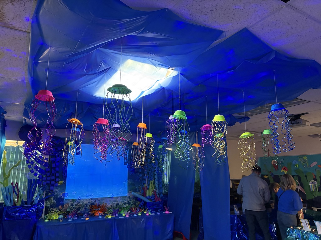 A classroom transformed into a large-scale ocean habitat, with blue fabric draped across the ceiling and handmade sea creatures hanging overhead, surrounding student-created animal models and adaptation projects.