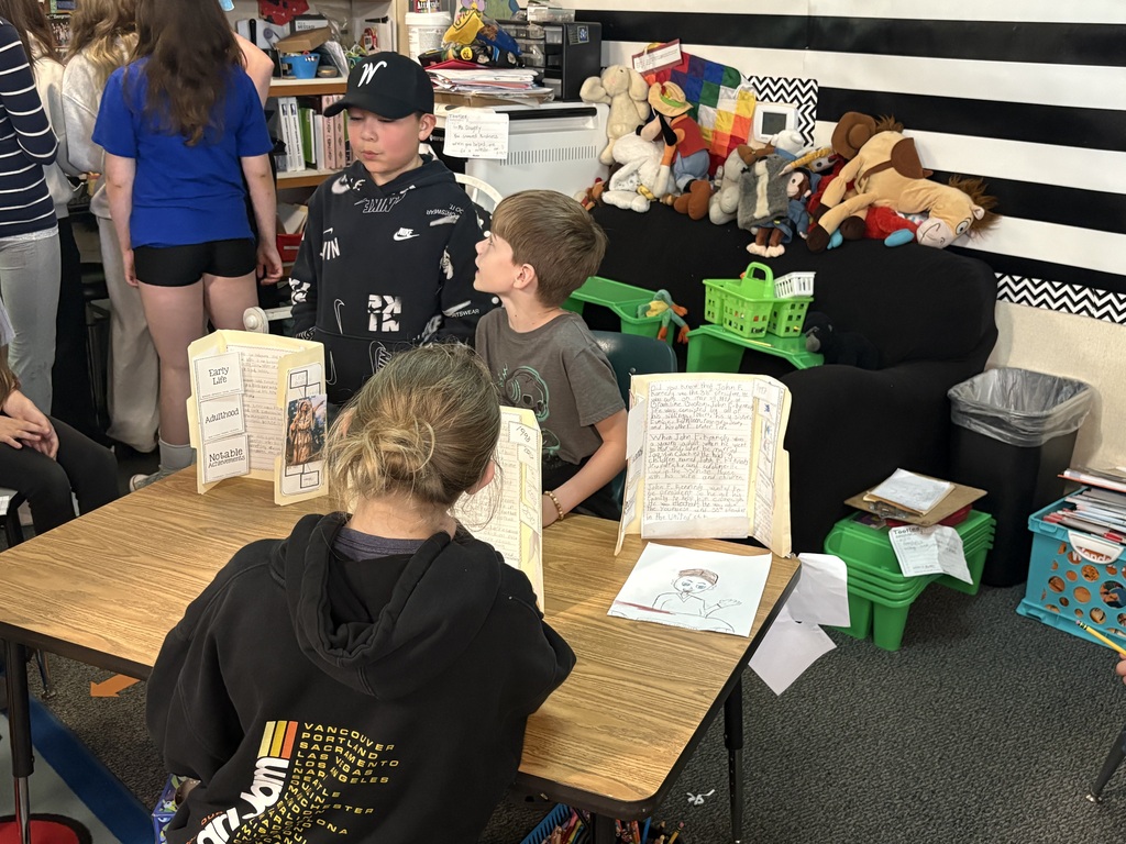 Third grade students at Evergreen STEM Elementary stand beside biography displays and presentation boards, sharing research projects about notable historical and contemporary figures with visitors during the school’s first Biography-palooza event.
