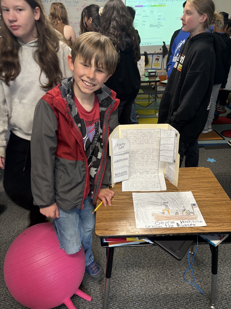 Third grade students at Evergreen STEM Elementary stand beside biography displays and presentation boards, sharing research projects about notable historical and contemporary figures with visitors during the school’s first Biography-palooza event.