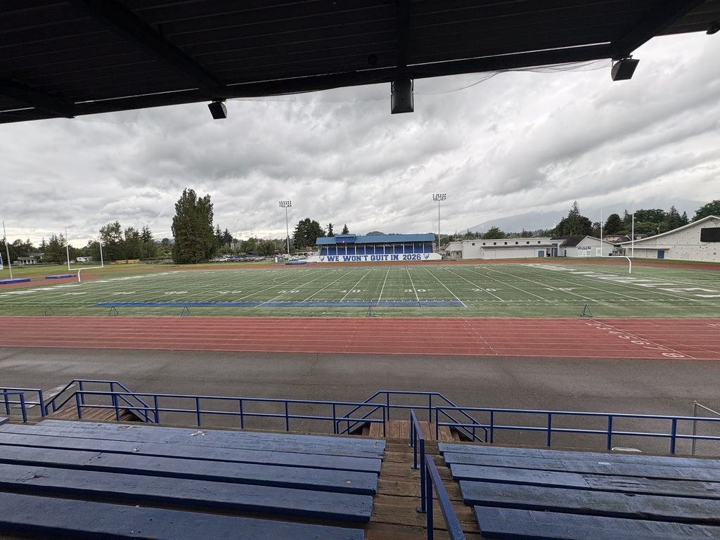 Wide view of the existing Sedro-Woolley High School stadium from the grandstands. Blue bleachers and railings frame the foreground, with the red running track and artificial turf football field stretching across the center of the image. The visitor stands displaying the message “WE WON’T QUIT IN 2026” are visible in the distance, along with stadium lights, school buildings, and trees. Dark, overcast clouds fill the sky above the facility.