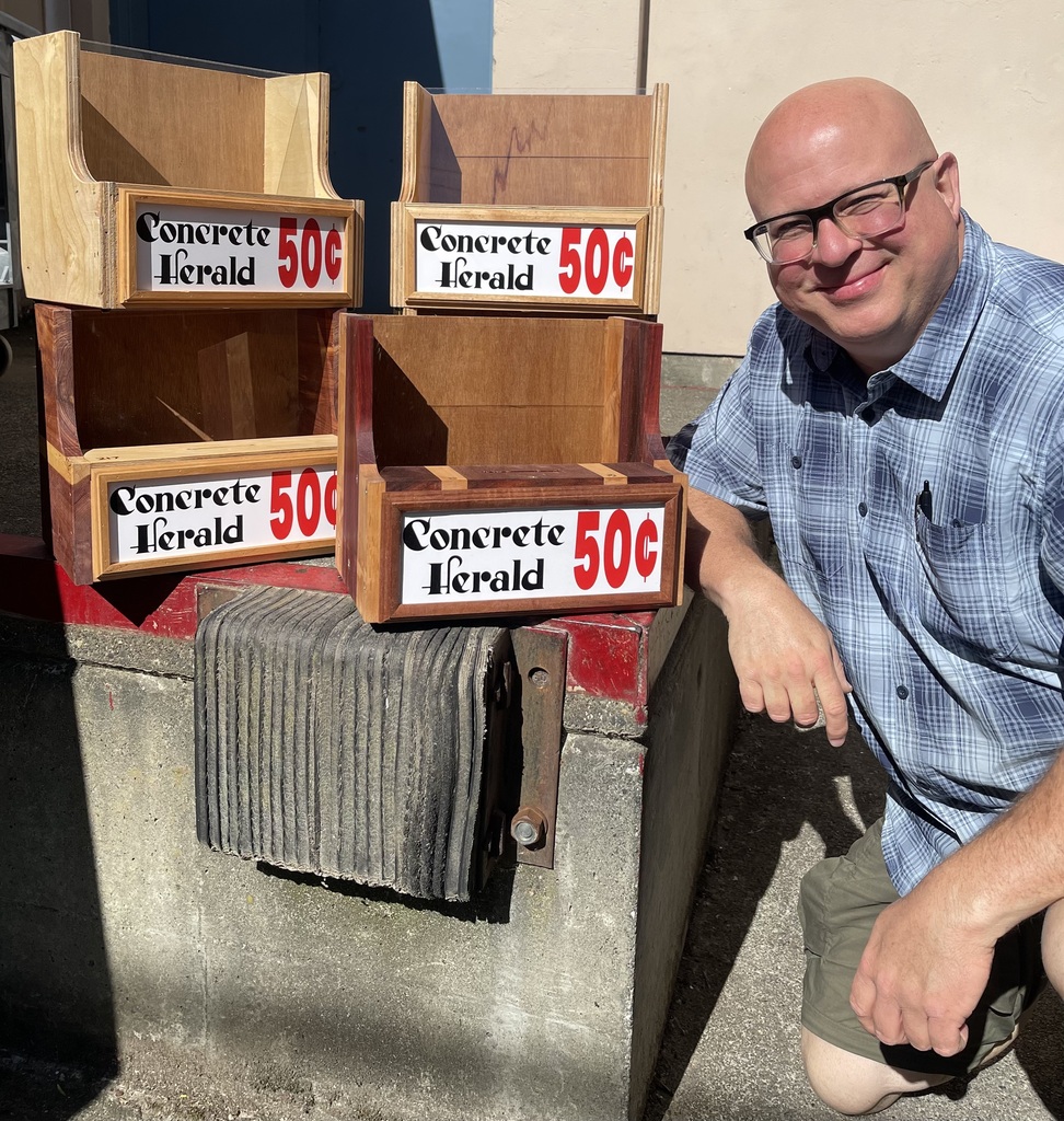 Jason Miller, editor and publisher of the Concrete Herald, kneels beside four handcrafted wooden countertop newspaper racks built by Sedro-Woolley High School Wood Production students. The racks display Concrete Herald signs with a 50-cent price label. The photo was taken outdoors in bright sunlight near a loading dock. The project is part of a partnership between the newspaper and the school's Career and Technical Education program, with Miller providing donations to support student learning.