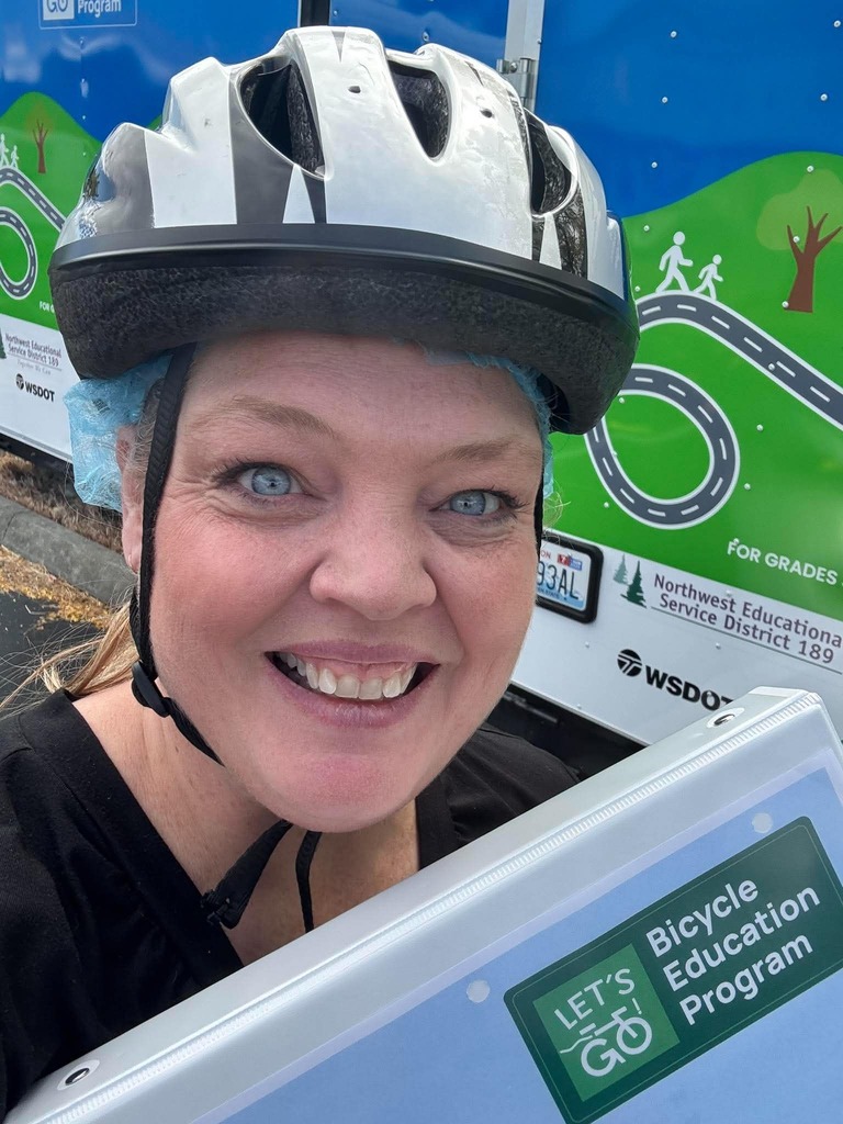 Evergreen PE Teacher Jennifer Batz smiles for a selfie while wearing a bicycle helmet and holding Bicycle Education Program training materials. A NWESD 189 Bicycle Education Program trailer is visible behind her.