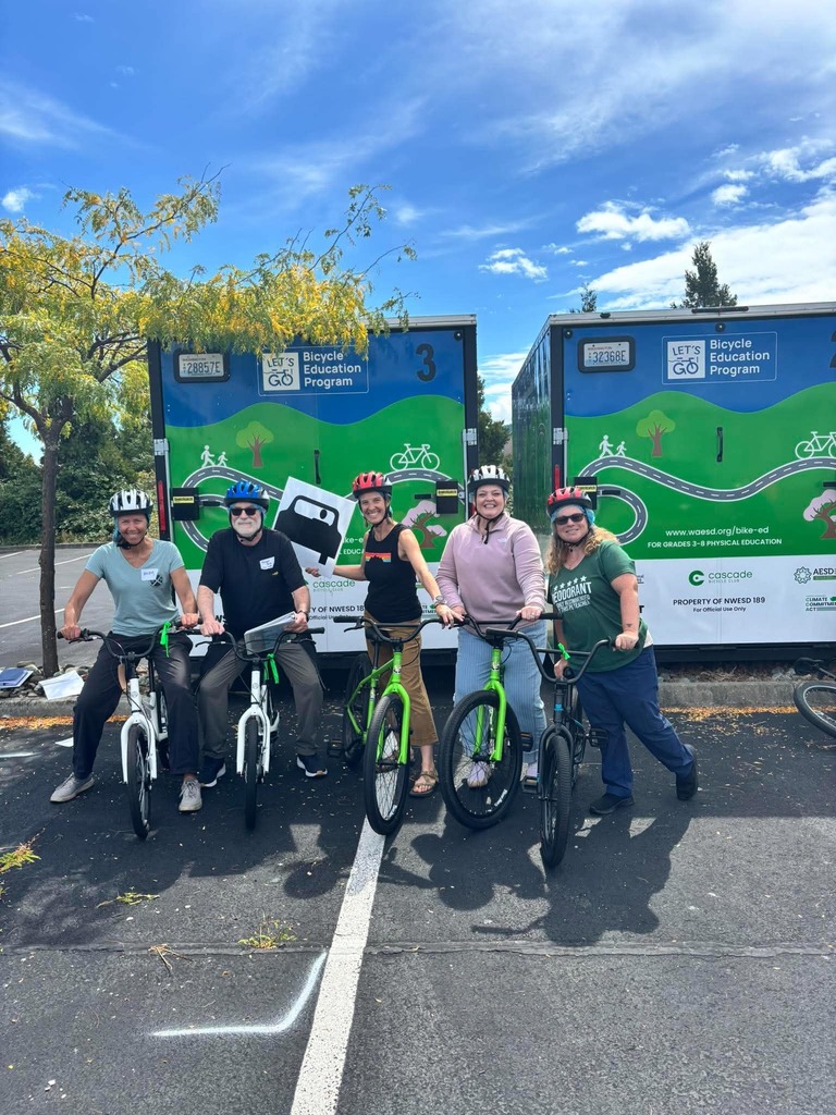 Five Sedro-Woolley School District staff members wearing bicycle helmets pose with bicycles in front of two colorful Bicycle Education Program equipment trailers during safety training at NWESD 189.