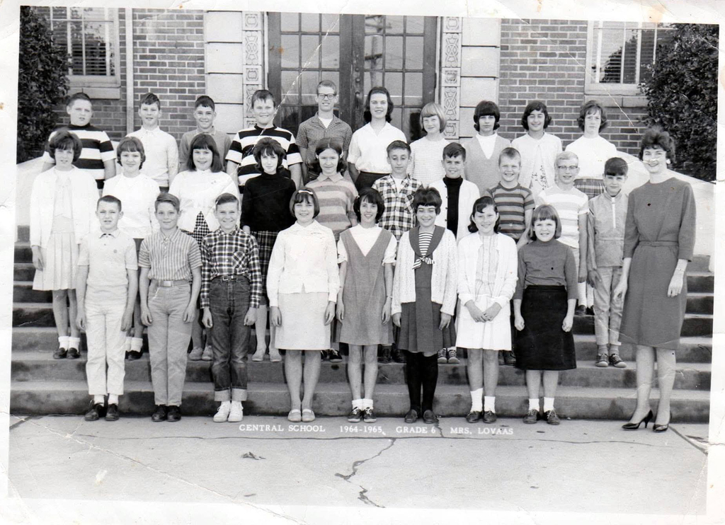Black-and-white class photograph of Central School's 1964-65 sixth-grade class with teacher Mrs. Lovaas. Students pose in three rows on the front steps of the brick school building. The photograph was shared by former student Patti Henderson Baines in celebration of Central Elementary's 100th anniversary.