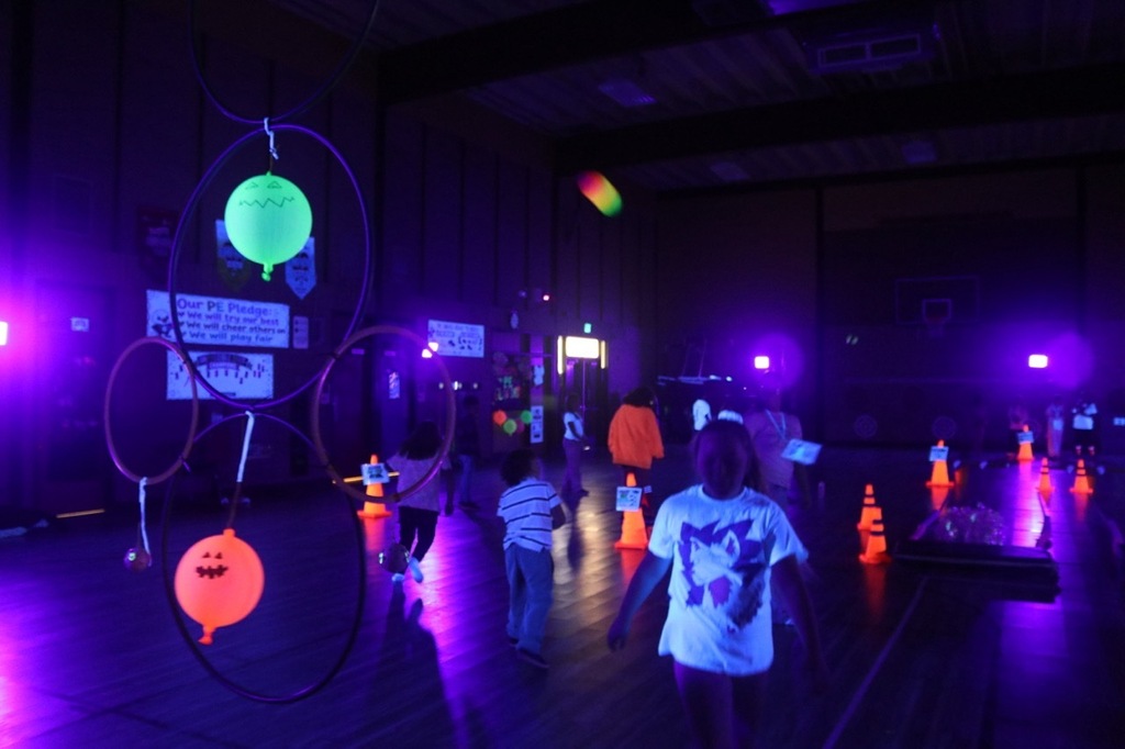 Friday Night Football: Students toss footballs through suspended hula hoops, aiming for glowing targets in the darkened gymnasium illuminated by black lights.