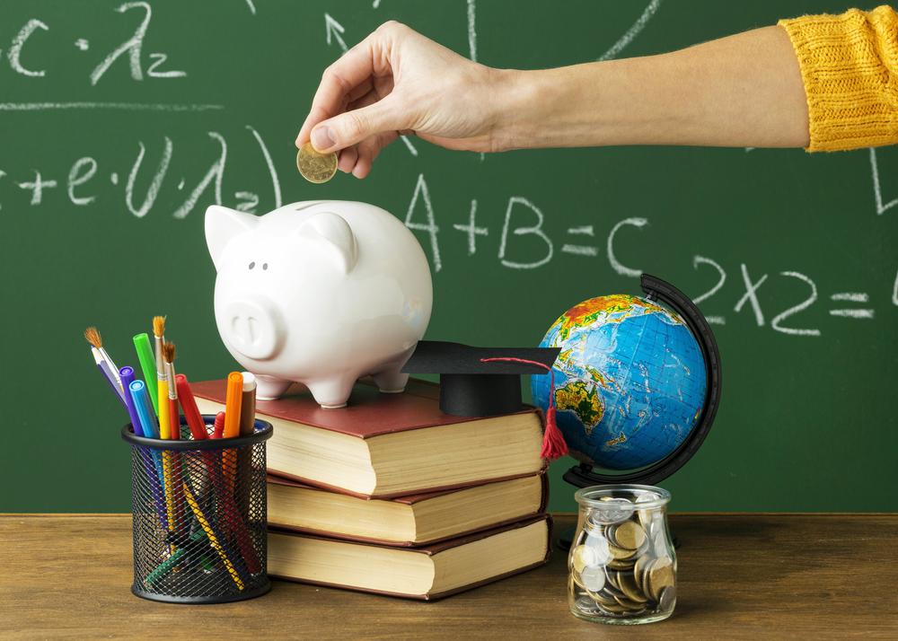 Image of someone putting a coin into a piggy bank on a stack of books, within a classroom.