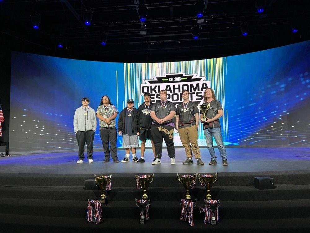 A group of six people stands on a stage with a vibrant blue and yellow background displaying "Oklahoma Esports." They hold trophies, conveying a triumphant mood. Four additional trophies are placed on the front of the stage. An American flag is visible to the left.
