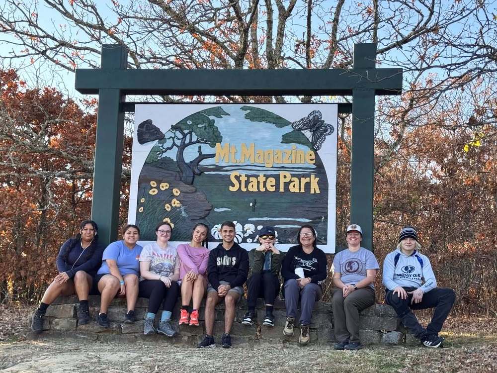 A group of eight people sits on a stone ledge in front of a "Mt. Magazine State Park" sign, smiling amidst autumn trees. The mood is cheerful and relaxed.