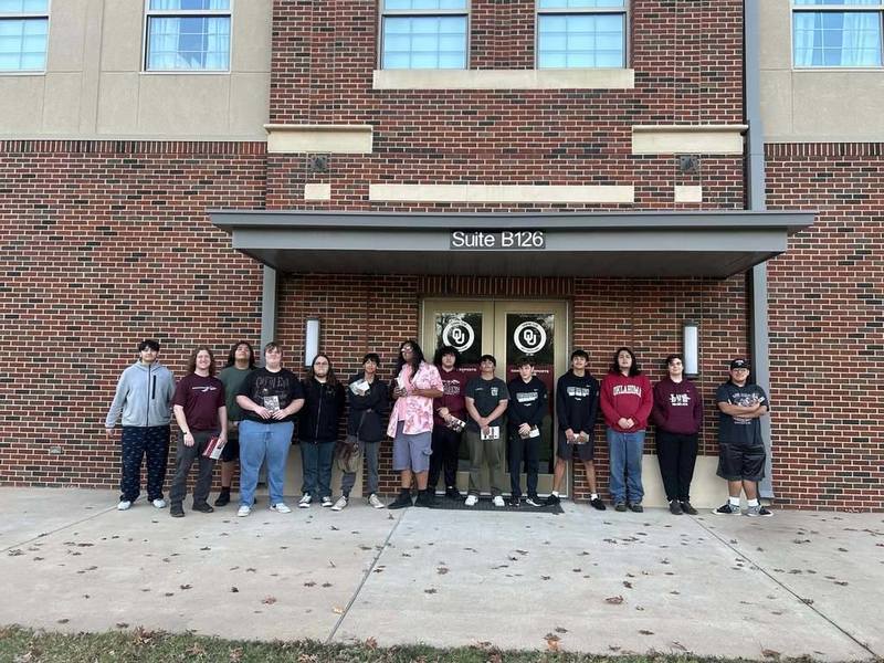 A group of people stands in front of a brick building with large windows, under a sign reading "Suite B126." They appear relaxed and happy.
