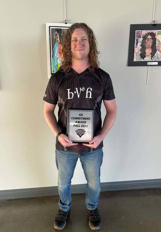 A person with long curly hair smiles while holding a "GG Commitment Award Fall 2025" plaque. They're wearing jeans and a dark shirt in a gallery setting.