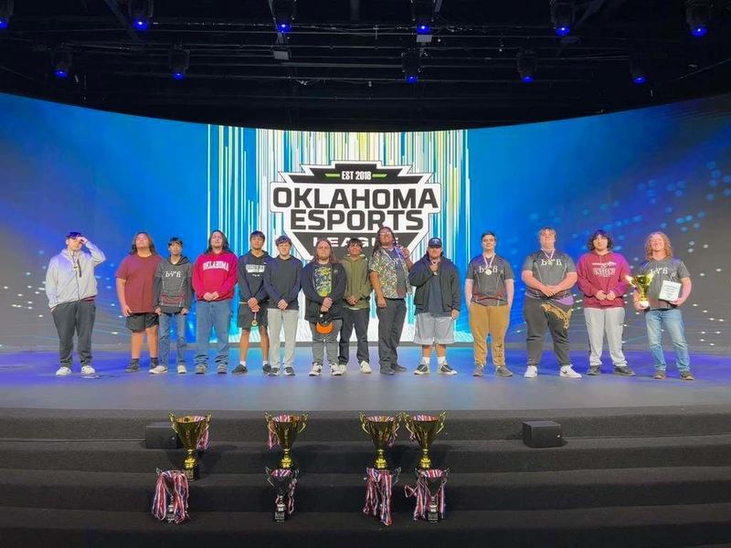 A group of people standing on a stage in front of an "Oklahoma Esports" banner, with trophies lined up in front. The mood is celebratory.