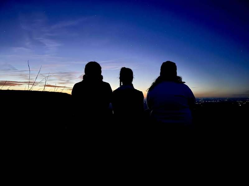 Silhouettes of three people sitting side by side, facing a vibrant twilight sky. The scene conveys a serene and contemplative mood.