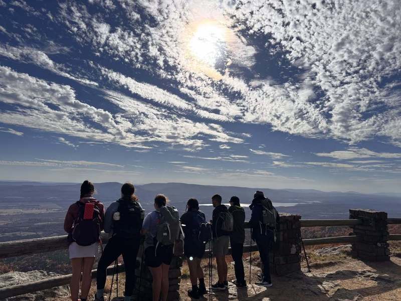 A group of hikers with backpacks stands at a rocky lookout, enjoying a vast scenic view under a bright sun and a sky filled with wispy clouds.